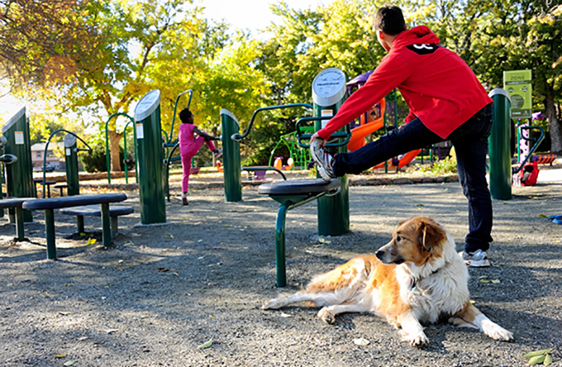 A dog relaxes near a young man stretching out at the new Fitness Zone at Silverman Park in Denver, CO. Health, exercise.
